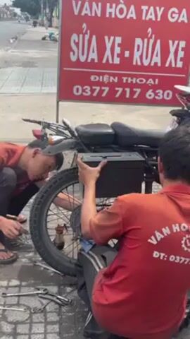 Two mechanics in red shirts are repairing the rear tire of a black motorcycle next to a red sign that advertises motorcycle repair and washing services with a phone number. One mechanic kneels and works on the tire while the other sits and holds a black box above the tire. Wrenches are scattered on the ground nearby. The scene is outdoors on a paved surface next to a road with passing traffic.