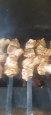 The video shows pieces of seasoned meat being grilled over hot coals. The meat is arranged on skewers and placed directly on the grill. The flames and smoke from the coals are visible, indicating that the meat is actively cooking. The lighting is dim, suggesting an outdoor or indoor cooking environment. The camera pans across the grilling meat, showing different angles and pieces.