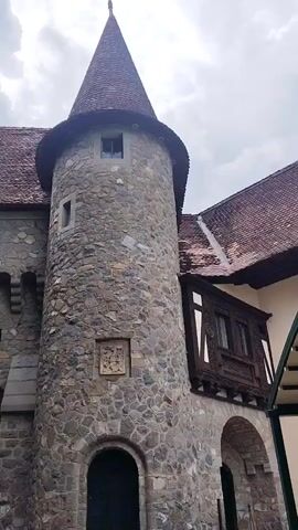 This video shows a stone castle tower with a conical roof. A group of people are walking towards an archway in the castle wall. One woman in the foreground is holding up a smartphone to take a picture of the castle. The sky is overcast.