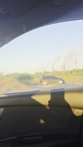 The video shows a view from inside a car, looking out the passenger window. The window is open, and the landscape outside is blurred due to the car's motion. In the distance, there are several tall structures with smoke rising from them, suggesting an industrial area. Trees and vegetation are visible along the side of what appears to be a road. The interior of the car is dark, with the armrest and door panel visible in the foreground. A shadow of a person is cast on the armrest. The sky is clear and bright, indicating daylight.
