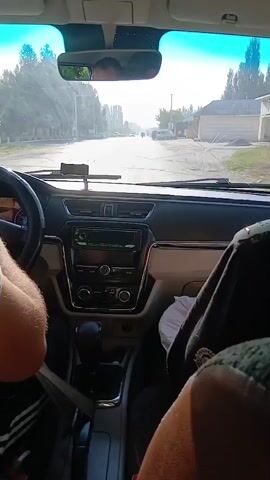 The video shows the interior of a car from the perspective of a passenger. The driver's hands are visible on the steering wheel. In the passenger seat, another person is mostly obscured by a dark blanket or clothing, with only their legs and one hand visible. The dashboard of the car, including the radio and gear shifter, is clearly visible. The car is driving on a paved road with trees and buildings lining the sides. The sun is shining brightly, creating glare on the windshield. In the distance, other vehicles and people can be seen on the road. One person is seen gesturing with their hand.