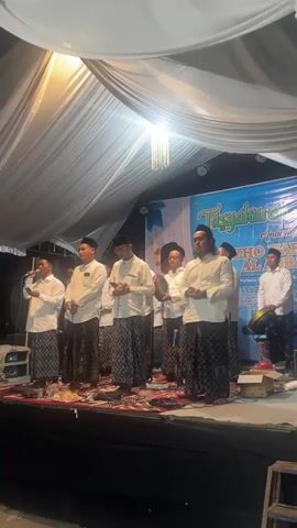 A group of approximately ten men in traditional Islamic attire, including white long-sleeved shirts and patterned sarongs, stand on a raised stage. Some hold microphones and appear to be singing or reciting. One man on the right plays a tambourine. Behind them, a blue banner with text that includes "TASYAKURAN" is visible. The stage is decorated with white fabric draped from above, and a chandelier-like light fixture hangs from the ceiling. Various items, including speakers, a microphone stand, and boxes, are present on the stage floor. The lighting is a mix of stage lighting and ambient light.