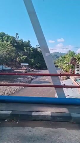The video is a first-person perspective shot from a bridge overlooking a river. The sky is clear and blue with a few scattered clouds. The river flows through a valley surrounded by lush green hills and trees. The riverbed is rocky and appears to be shallow in some areas. There are some structures visible along the riverbanks, including a small building and what appear to be market stalls or roadside shops. Some people are visible near the river, possibly bathing or engaging in other activities. The bridge railing, painted blue and red, is in the foreground. The camera pans slightly, offering a dynamic view of the landscape. The overall impression is a serene and natural environment with signs of human activity.
