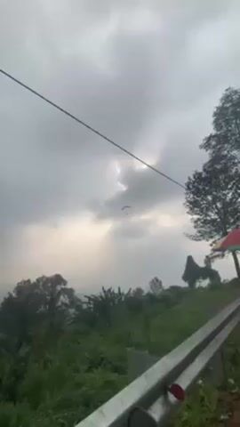 The video shows a person paragliding against a cloudy sky. The landscape below is green with trees and vegetation. A metal guardrail is visible in the foreground.