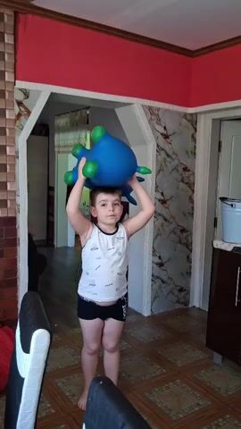 A boy is playing with a blue inflatable toy. He is in a room that appears to be a kitchen. The boy is wearing a white tank top and black shorts. He is holding the toy over his head at the beginning of the video. He then throws the toy and jumps, and then lands on the toy.