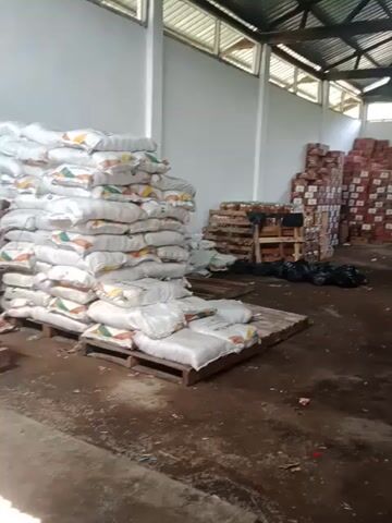 The video shows the interior of a warehouse.  Stacks of boxes and pallets laden with sacks are stored inside.  Several individuals are gathered around a small truck.  They are holding a banner, although the banner is blurry and not completely legible.  The floor appears to be dirty.  The light appears to come from an opening in the roof.