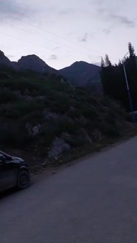 A black car is parked on the side of a paved road. The road winds through a mountainous area with sparse vegetation. The sky is overcast, and it appears to be dusk or dawn. In the distance, a small village is visible nestled in a valley. There is a utility pole with wires and a street lamp along the road. There are also some construction materials and a wooden bench visible.