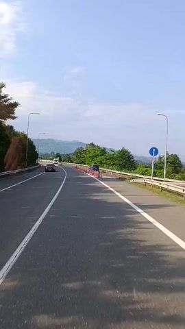 A car is driving down a highway. The road is paved and has white lines marking the lanes. There are trees and mountains in the background. The sky is blue with some clouds. Several other cars are visible on the road. There are orange traffic cones lining the side of the road.