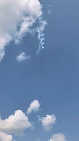 The video shows a clear blue sky with some fluffy white clouds. In the upper part of the frame, a thin trail of white smoke is visible, twisting and turning as it descends. At the end of the smoke trail, a small, bright object appears to be falling. The object is small and difficult to discern, but it seems to have a light-colored body and possibly a dark tip. The smoke trail suggests that the object may have been propelled upwards at some point. The video is taken from a low angle, looking up at the sky.