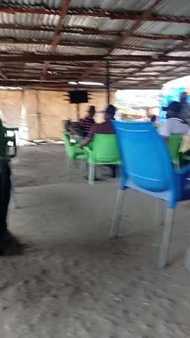 The video shows the interior of an open-air structure with a corrugated metal roof. Several groups of people are seated at tables, some watching a television. The tables are surrounded by plastic chairs of various colors, including green and blue. The ground is dirt. One person walks across the space between the tables. There are empty beer bottles on one of the tables.