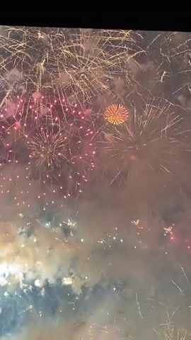 The video captures a fireworks display. The camera angle is looking upwards at the sky, filled with bursts of colorful fireworks, smoke, and light. In the foreground, there are trails of light and smoke, creating a layered effect. The fireworks vary in color, with prominent hues of gold, pink, white, and green. The display appears to be ongoing, with multiple explosions and fading embers visible. There are no people or identifiable objects in the scene, other than the fireworks themselves. The environment is dark, presumably nighttime, which accentuates the brilliance of the fireworks.