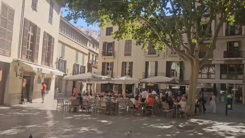 The video pans from a bustling outdoor cafe with numerous patrons seated at tables to a street scene with buildings, trees, and people. A small red car is visible, as is a utility vehicle with a large bin. The camera then focuses on a large, ornate church with Gothic architectural features, framed by trees. The sky is clear and blue, indicating a sunny day.