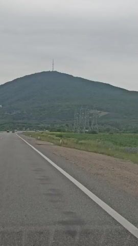 The video shows a landscape from a moving vehicle. A paved road with a white line runs along the bottom of the frame. Beyond the road is a grassy field with some tall plants and scattered purple flowers. In the background, there are rolling hills covered in trees. A large electricity pylon stands in the middle ground, and a mountain peak is visible in the far distance. The sky is overcast.