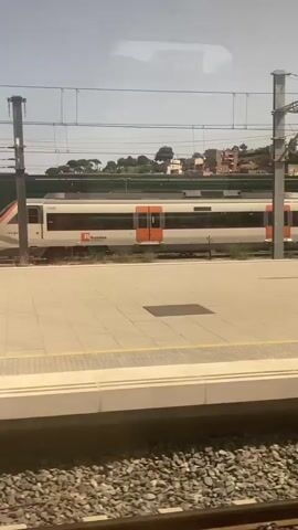 The video shows a train at a station. The train is white with orange doors and purple accents. The station platform is paved with light-colored tiles and has a yellow line along the edge. There are poles and overhead wires for the train. In the background, there are buildings and trees on a hill. The video is a still shot and does not show any movement or people.