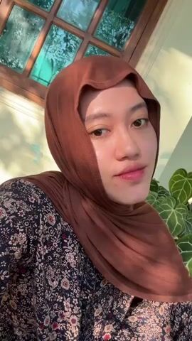 A young woman with a brown hijab and a dark floral patterned shirt is centered in a medium close-up, looking directly at the camera. She has dental braces on her upper teeth. Her expression transitions from a slight smile to a broad smile. A multi-pane wooden window is visible in the background to her left, and green plants are to her right, all illuminated by natural light.