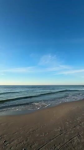 This is a vertical, full-shot video of a beach and the ocean under a clear blue sky with some wispy clouds. The sand is damp and shows footprints. Gentle waves roll onto the shore. In the distance, a line of wooden posts protrudes from the water. The sun is low in the sky, casting a warm light on the scene.