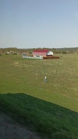The video shows a rural landscape, likely filmed from a moving vehicle. The foreground consists of grassy fields and a paved road. In the distance, there's a small village with houses having red roofs. The sky is clear and blue. A road sign depicting a bus is also visible. The overall environment seems peaceful and typical of a countryside area.