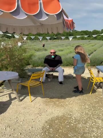 A man with a shaved head is sitting on a bench under an umbrella, in front of a lavender field. He is wearing a black t-shirt, white pants, and black shoes. He has tattoos on his arms and is wearing sunglasses with a green flower-shaped accessory on his shirt. A small dog is sitting on the ground in front of him. A young girl is standing to his right, holding a phone and taking a picture of him. In the background, there are rows of lavender plants and some white tents. There is also a white table and yellow chairs in the foreground.
