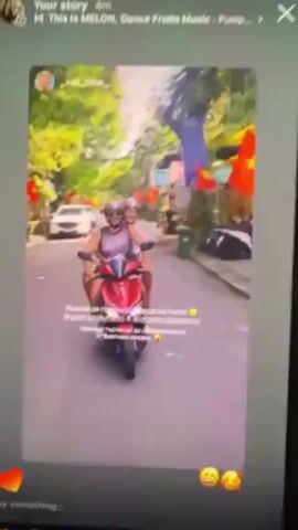 A video shows two people riding a red scooter on a narrow street lined with buildings and Vietnamese flags. The person driving the scooter is wearing a light-colored tank top, sunglasses, and a helmet. The passenger behind them is also wearing a helmet and a light-colored outfit. A white car is visible parked on the side of the road. The video appears to be a screen recording of a social media post. The text in the post is in Russian. The background music is a dance song.