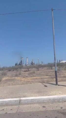 The video shows a large industrial complex, likely an oil refinery, under a clear blue sky. The complex is composed of many interconnected structures including towers, pipes, and storage tanks. Several flares burn with orange flames and produce black smoke. The complex sits on a slightly elevated, arid landscape with sparse vegetation. The camera appears to be moving, possibly from a vehicle on a nearby road. The foreground shows a portion of the road. The overall scene conveys a sense of industrial activity.