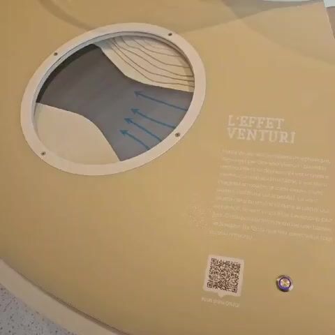 Here is a detailed description of the video:

The video shows a diagram explaining the Venturi effect. The diagram is circular with a white frame and clear cover, mounted on a tan surface. Inside the circle, there is a visual representation of airflow through a constricted space, showing arrows indicating acceleration. 

Next to the diagram is text, written in French, explaining the Venturi effect, detailing how air accelerates when it passes through a narrower space, resulting in a decrease in pressure. A QR code and a button labeled "PLUS D'INFOS ICI" are present beneath the text. The button is pushed at the beginning of the video.