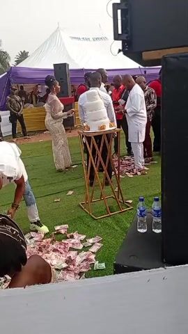 The video shows an outdoor event, possibly a wedding celebration, where money is scattered on the ground. A woman in a shimmering dress stands near a tiered cake on a gold stand. Several men are standing around the cake. One man is bending over, apparently gathering the money from the ground. There are several other people in the background, and a white tent is visible. Speakers are also present, and there are bottles of water on a black surface. The ground appears to be artificial turf.