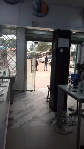 The video shows the interior of a store, possibly a phone shop, from a stationary position looking towards the entrance. The floor is tiled with gray marble pattern. In the foreground, there are display tables with various items that look like phones and accessories.  The open glass door reveals a street scene with two people standing outside, cars parked along the roadside, and other buildings in the background.  A tall, dark cabinet stands near the door inside the store.  In the right foreground, there is a white table with a fan and other items on it.  Hanging from the ceiling are promotional signs for "Infinix" phones. Light streams into the store from the open doorway, illuminating the interior and casting shadows on the floor. The general environment appears to be a bustling commercial area.