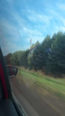 The video shows a view from inside a moving vehicle looking out the passenger window. The landscape outside consists of a dense forest of green pine trees on a sloping hill. A tall telecommunications tower is visible above the trees on the far right side of the frame. The sky is blue with wispy white clouds. The road is partially visible in the foreground, appearing as a blurred strip of gray and white lines, indicating the vehicle is in motion. A side view mirror is visible on the left side of the frame. The overall impression is a scenic drive through a wooded area.