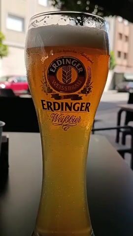 The video shows a close-up, slightly angled shot of a tall glass of Erdinger Weissbier, a German wheat beer. The glass is frosted with condensation, indicating it is cold. The beer itself is a cloudy golden-amber color with a thick white head of foam at the top. The Erdinger Weissbräu logo is prominently displayed on the glass. The background is blurred but appears to be an outdoor setting, possibly a restaurant or cafe patio, with tables and chairs visible, as well as a red car and building in the distance. The lighting suggests it is daytime. There are no people visible in the frame. The video appears to be a static shot of the beer, with no discernible action or movement.