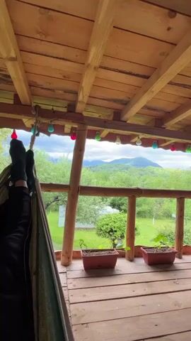 From a shaded wooden porch, a person lounges in a hammock, feet clad in black socks visible in the lower left corner of the frame. Overhead, string lights of varying colors hang from the wooden ceiling. The porch overlooks a lush green landscape, featuring a yard and a distant view of tree-covered mountains under a cloudy sky. There are two rectangular flower pots on the porch with small plants.