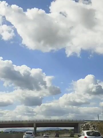 A car drives on a highway under a bridge, with clouds and mountains in the background. The sky is blue with white clouds. The bridge is made of concrete and has a railing. Several cars are on the highway.