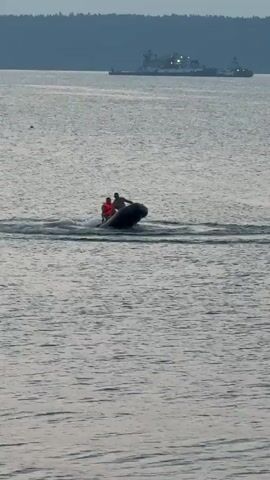 Two people are in a small inflatable boat on the water. One person is wearing an orange life jacket. The boat is moving and creating a wake. In the background, a larger ship is visible on the horizon. The sky is overcast and the water is choppy.
