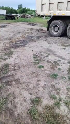 A muddy, rutted dirt path stretches across the frame, with puddles of dark, oily-looking water scattered across its surface. In the background, a large, white dump truck is parked, its rear wheels and tires visible. Beyond the truck, a sparse, rural landscape unfolds under a cloudy sky. Several simple, weathered buildings, some white, others appearing derelict, are scattered amongst patches of green vegetation and trees. A utility pole with wires is visible in the distance, and what appears to be another trailer or vehicle is partially obscured on the left side of the frame. The overall impression is one of a working, perhaps agricultural or industrial, outdoor space in a rural setting.