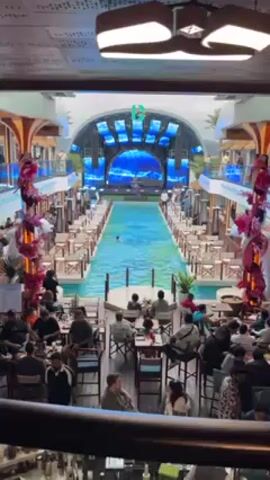 The video shows the interior of a large restaurant that has a swimming pool running down the middle of the dining area. There are tables set up on either side of the pool where people are sitting and eating. Some people are standing around as well, near the bar. At the far end of the pool is a stage area with large screens showing animated ocean scenes. On the second level of the restaurant, people are visible looking down at the scene. The lighting in the restaurant is modern, and the decor includes tall floral arrangements. There appears to be a party or event happening.