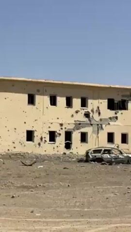 The video shows a damaged two-story building with numerous bullet holes and structural damage, a wrecked car in front of it, and several people walking around the site. The building is painted in a light beige or yellowish color. The facade has several visible bullet holes and areas where the concrete is broken away. The roofline is flat with a slight overhang. All the windows are either broken or missing. 

In front of the building, there is a severely damaged white car, possibly due to an explosion or heavy impact. The vehicle is surrounded by debris and rubble. The ground is dry and barren, appearing to be a dirt or gravel surface. Debris such as broken metal and building fragments are scattered across the ground.

There are several people walking around the area, dressed in civilian clothes. They appear to be inspecting the damage. Further in the background, what looks like a wire fence surrounds the perimeter. The sky is clear and blue, suggesting a sunny day. There are hills or mountains in the distant background.