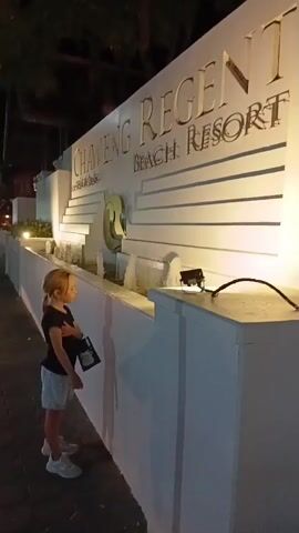 The video shows a young girl standing near a large, illuminated sign for the "CHAWENG REGENT BEACH RESORT". The sign is white with large, gold, embossed lettering and a decorative element that resembles a stylized "G". Below the sign, a water feature with several spouts is visible. The girl, wearing a black t-shirt and light-colored shorts, stands on a paved walkway. She holds a dark-colored bag with white lettering. The scene appears to be at night, as indicated by the artificial lighting illuminating the sign and the darkness in the background. The weather seems pleasant as there are people walking in the background and the girl is dressed in short sleeves and shorts. The overall mood appears calm and relaxed. The location is clearly identified by the resort's sign as Chaweng Beach, Thailand.