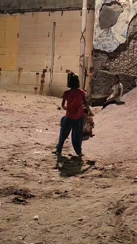 A video shows two young children playing in a sandy, uneven area with rubble and a concrete wall in the background. A street light illuminates the scene. One child, wearing a red shirt and blue pants, is in motion, possibly dancing or running. Another child, in lighter-colored clothing, sits on the ground. A third, older child sits further back against the wall. The environment appears somewhat neglected, with exposed rebar and graffiti on the wall.