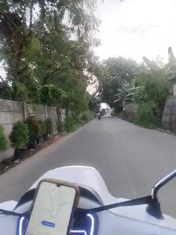 The video shows a first-person perspective of someone riding a white scooter or motorcycle on a paved road. A smartphone is mounted on the handlebars, displaying a navigation app with a green route. On the left side of the road, there is a low concrete wall with overgrown grass and vegetation. Further down the road, there are trees and some buildings or structures. Other vehicles, including motorcycles, can be seen in the distance. The sky appears to be overcast or at dusk, with a soft light filtering through the trees. The overall environment suggests a rural or semi-urban setting.
