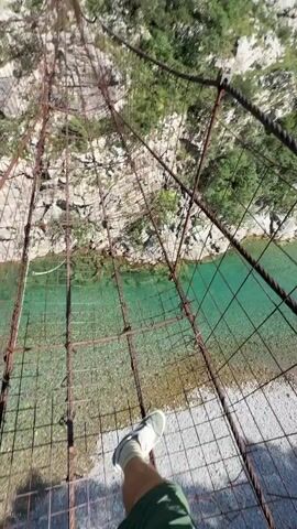 A person stands on a metal suspension bridge above a river. The bridge is made of wire mesh and has thick wire cables running along its sides. Below the bridge, a clear, turquoise river flows over a rocky bed, with some sections having a lighter, more gravelly appearance. The river is surrounded by lush green mountains on both sides, with some bare rock faces visible. The sky is bright blue with a few faint wisps of clouds. The camera moves, showcasing the river from different angles, and then captures the person's feet on the bridge as they walk.