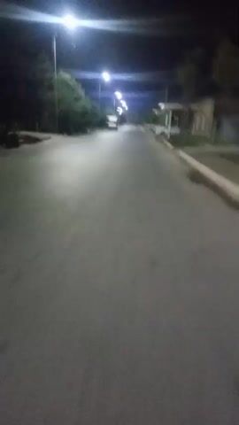 The video shows a dimly lit paved road at night, illuminated by streetlights. Trees line the left side of the road, while buildings and parked cars are visible on the right. The camera appears to be moving forward, possibly from a vehicle or a person walking, as indicated by the slight shake and the perspective. The overall atmosphere is quiet and still, with no visible human activity besides the implied movement of the camera.
