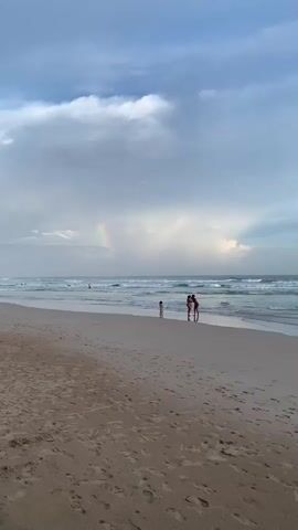 A person in a striped shirt stands on a sandy beach facing the ocean. Two other people are further down the beach near the water. There are waves in the ocean and a rainbow in the sky. The sky is cloudy.