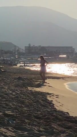 A person is standing on a sandy beach near the water. The sun is shining brightly, creating a glare on the water. In the background, there are buildings and mountains. The person appears to be wearing a light-colored outfit and is facing away from the camera, towards the water.