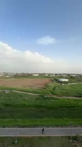The video presents a wide, static shot of a rural landscape under a bright blue sky with scattered white clouds. In the foreground, a paved path stretches horizontally across the frame, flanked by evenly spaced streetlights. Two individuals are visible on the path, both seemingly skateboarding or riding scooters. The landscape transitions from the path into expansive green fields, with a large patch of brown earth breaking up the verdant expanse. In the distant background, a cluster of buildings and structures are vaguely discernible against the horizon. The overall ambiance of the scene is peaceful and open.