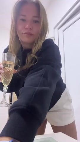 A young woman with long, light brown hair, wearing a black sweatshirt and white shorts, holds a champagne flute filled with a sparkling beverage. She has light-colored nail polish and a smartwatch on her left wrist. She is smiling and dancing or swaying to music, appearing happy and celebratory. The background shows white walls, a light-colored wooden cabinet, and a white door, suggesting an indoor setting like a home. The lighting is bright. The video is set in Sotonići, Montenegro.