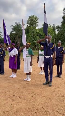 A medium shot shows nine young people dressed in various uniforms standing in formation on a dirt field under a cloudy sky. They are holding flags of different colors, and several individuals are saluting. From left to right, the flags are green, purple, white, and black. Trees and greenery are visible in the background.
