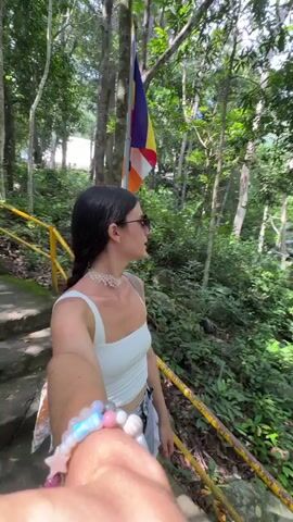 A first-person perspective video shows a woman with dark hair and sunglasses wearing a white top and shorts. She is holding a phone out, likely recording a selfie video, as she walks up a stone staircase with a yellow handrail. To her left, there is a flag with multiple colors, including purple, yellow, orange, and white, attached to a pole. The background is a lush green forest with tall trees and foliage. The sounds of birds chirping and rustling leaves can be heard.