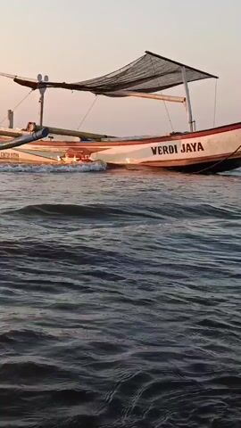 The video shows a traditional Indonesian fishing boat named "VERDI JAYA" bobbing on the water near a beach at sunset. The boat is painted orange, white, and red, with a canopy providing shade. The water is choppy, with small waves breaking against the hull of the boat. The sky is a pale orange, indicating the sun is low on the horizon. The scene is peaceful and serene.