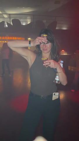 A woman in a dimly lit room holds two small glasses up to her eyes. She has dark hair and is wearing black pants, a brown top, a watch, bracelets, and rings.  She has a water bottle tucked under her left arm. A cell phone is visible in her right pants pocket. Other blurry people are visible in the background. A wooden floor is present.  There appear to be decorative elements on a balcony behind her. Red lights illuminate the scene.
