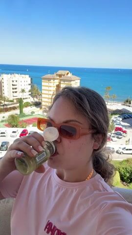 A woman is drinking from a glass bottle. She is wearing a pink t-shirt with the word "AMOUR" written on it in red, orange-tinted sunglasses, and her hair is pulled back into a low ponytail. She is standing on a balcony overlooking a beach town with multi-story buildings, palm trees, a tennis court, parking lot, and a blue ocean under a clear blue sky. The label on the glass bottle is partially obscured but the word "GENJIBRE" is visible.