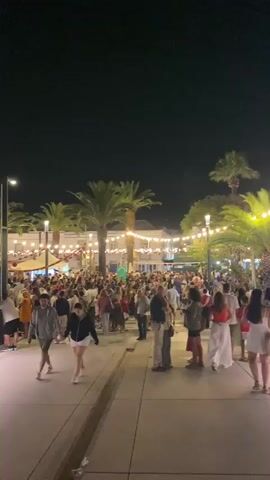 A nighttime outdoor gathering is shown. The scene is illuminated by strings of hanging lights and the ambient light from buildings. Several palm trees are visible, adding to the tropical or Mediterranean ambiance. A large crowd of people, dressed in casual summer attire, is spread across a paved area. Many individuals are walking, talking, and standing in groups, suggesting a lively event. In the foreground, two individuals are walking towards the camera, and others are visible further back. The overall atmosphere appears festive and social.